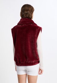 Gilet senza maniche color bordeaux realizzato in materiale morbido e peloso, con colletto alto e orlo elastico. Tessuto liscio con una tinta unita.