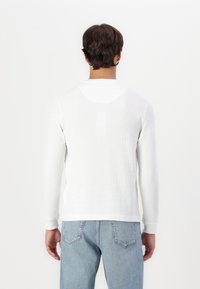 Weißes Langarmshirt aus strukturiertem Strickstoff mit Rundhalsausschnitt, kombiniert mit hellblauen Jeans, von hinten gezeigt.