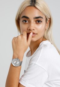 Montre analogique en argent avec un cadran rond, un bracelet en maille, deux sous-cadrans et un cadran gris texturé. Portée sur une main à ton de peau clair.