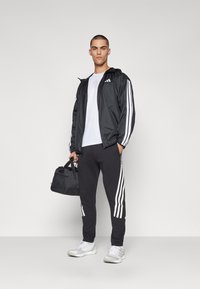 Sort windbreaker med hvide striber, parret med sorte sweatpants med matchende striber. Hvid t-shirt og grå sneakers fuldender outfittet.