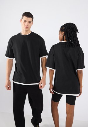 T-shirts noirs surdimensionnés avec des bordures blanches au col et aux manches ; portés par deux personnes, mettant en avant une coupe décontractée et une texture douce.