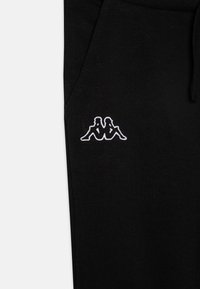 Črne trenirke iz mehkega blaga, z izvezenim belim logotipom dveh figur na levem stegenu. Prilagodljiv pas z vrvico.