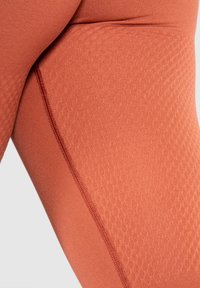 Strak zittende leggings in een gestructureerde roestkleur met een gladde afwerking. Kenmerkt zich door een naad die aan de zijkant loopt voor extra detail.
