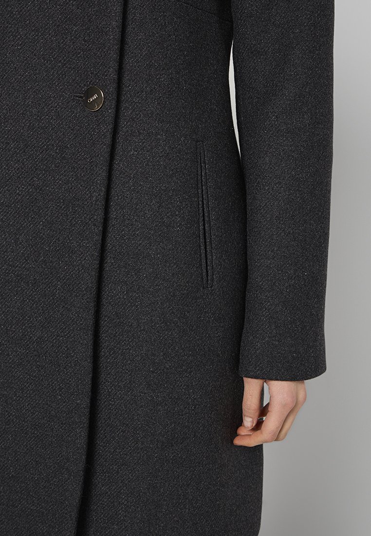 Manica di un cappotto di lana grigio scuro con un singolo bottone metallico e tasca a fessura, mano rilassata di lato contro uno sfondo neutro.