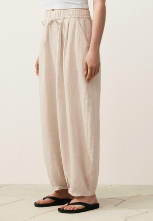 Žena oblečená v voľných béžových nohaviciach so šnúrkou a vreckami a v čiernych žabkách, stojaca na dláždenej podlahe pred jednoduchým pozadím.