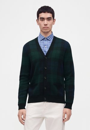 Homme portant un cardigan à boutons à carreaux vert foncé et bleu marine par-dessus une chemise à carreaux bleue et un pantalon blanc sur un fond uni.