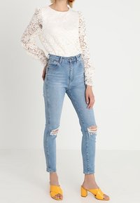 Blusa bianca in pizzo con maniche lunghe, abbinata a jeans skinny strappati di un blu chiaro e sandali con tacco gialli.
