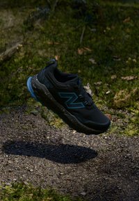 Chaussure de sport noire avec des accents bleus, tige texturée, semelle mouchetée et design robuste. Flottant au-dessus d'un sol naturel et herbeux.