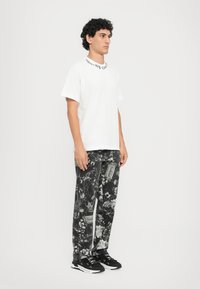 Balta medvilninė marškinėliai su juodu užrašu ant kaklo, derinami su laisvais juodais kelnais, turinčiais gėlių monohrominį raštą, ir juodais sportbačiais.