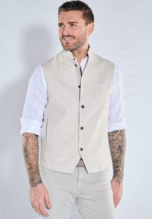 Beige sleeveless vest with a textured fabric, featuring a stand-up collar, five black buttons, and side pockets, worn over a white shirt.