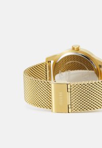 Goldmesh-Armbanduhr mit rundem Gehäuse, transparentem Rückdeckel und einemverschluss mit dem Markenlogo "GUESS." Texturiertes Armband und glänzende Oberfläche.