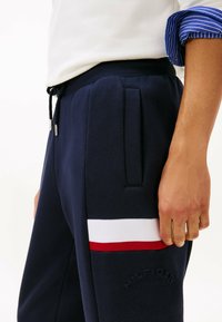 Marineblaue Sweatpants mit elastischem Bund, Kordelzug, seitlichen Taschen und farblich abgesetzten Akzenten in Weiß und Rot. "HILFIGER"-Logo auf der Seite eingeprägt.