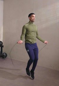 Sportisks vīrietis, ģērbies pieguļošā olīvzaļā zip-up džemperī un tumši zilos legingos, lecam ar melno lecamauklu sporta zālē.