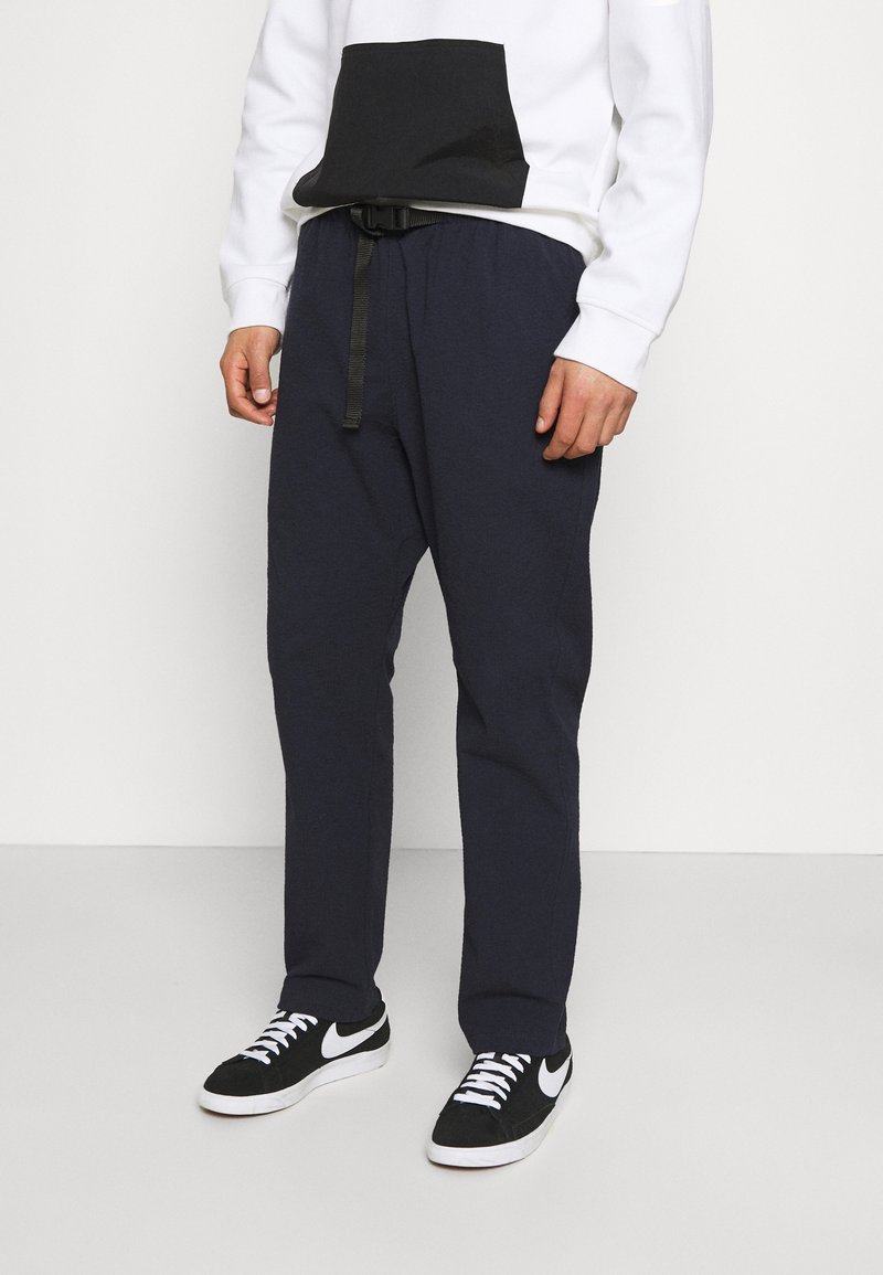 Námořnické kalhoty s elastickým pasem a černým pásem, kombinované s bílou mikinou s černou kapsou. Černé tenisky dotvářejí vzhled.