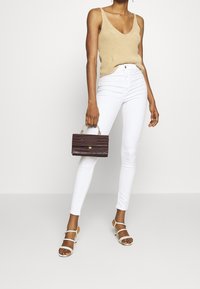 Top de tirantes de punto beige, jeans skinny blancos y sandalias de tacón con tiras en marrón claro. Un pequeño bolso texturizado marrón oscuro con herrajes dorados.