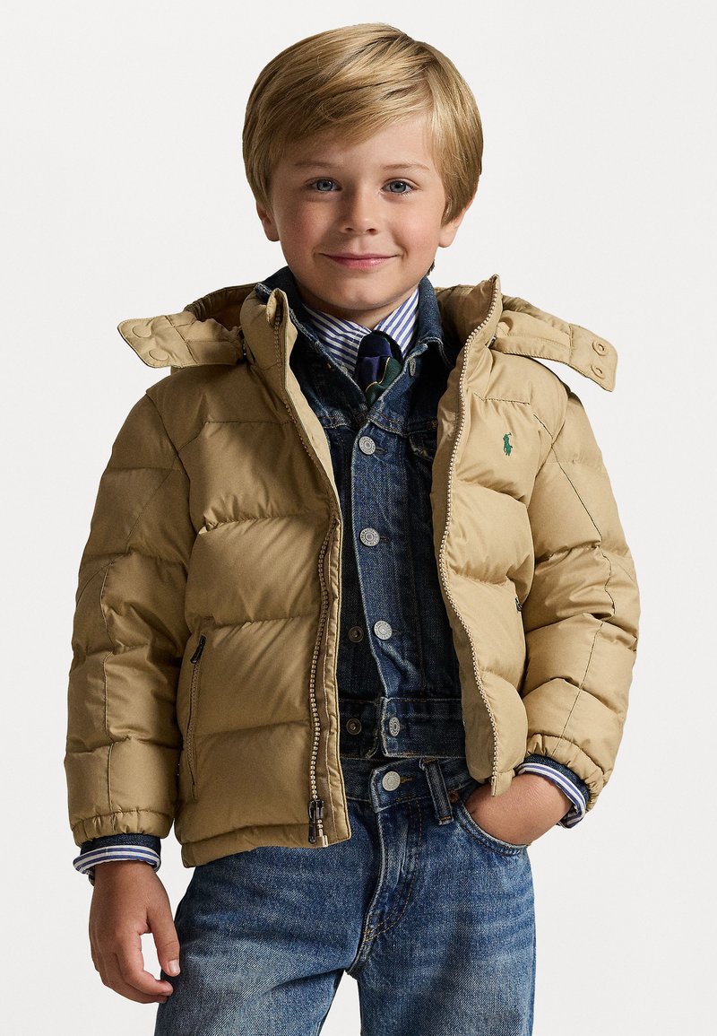 Μπεζ μπουφάν με κουκούλα, με ραμμένα πάνελ και ένα μικρό πράσινο λογότυπο, φορεμένο πάνω από πουκάμισο τζιν και μπλε τζιν.