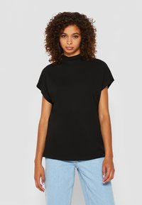 T-shirt noir à manches courtes avec un col haut, texture lisse et coupe décontractée, associé à un jean bleu clair. Aucun motif ni élément métallique visible.