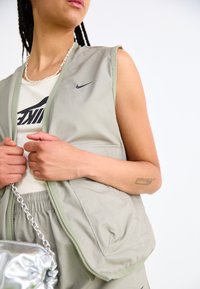 Světlé zelené bezrukávové tričko se zipem na přední straně a dvěma bočními kapsami, nošené přes bílé tílko s černým logem Nike.