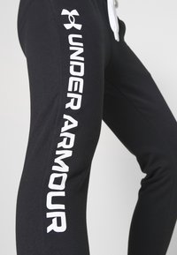 Svarta sweatpants i bomull med slim fit. Har en vit "Under Armour"-logotyp tryckt vertikalt längs ett ben.