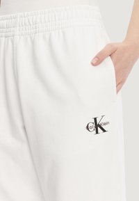 Bílé teplákové kalhoty vyrobené z měkké látky s elastickým pasem. Mají boční kapsu a černý detail s logem "Calvin Klein".