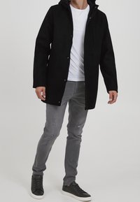 Mand iført en sort hættejakke, hvid t-shirt, grå slidte jeans og sorte sneakers, står på en ensfarvet baggrund.