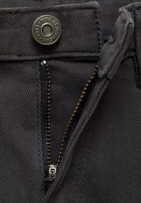 Tmavo šedá džínovina odhaľuje kovový gombík, kontrastný zips so zlatými zubami a detaily šitia podél zipsu.