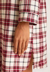 Tessuto a quadri rosso e bianco con accenti neri. Presenta un colletto, dettagli sui polsini e uno spacco laterale. Primo piano di una mano appoggiata sul tessuto.