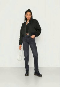 Jovem mulher com cabelo trançado, vestindo uma jaqueta preta curta, um top preto, jeans escuros e botas pretas, segurando uma bolsa branca.