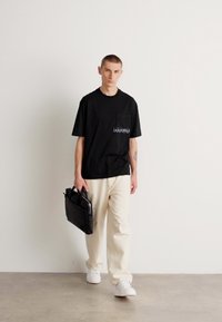 T-shirt negru din bumbac cu logo-ul "HUGO", asortat cu pantaloni largi de culoare crem și adidași albi, ținând o geantă din piele neagră.