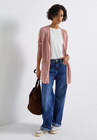 Un cardigan tricoté rose pâle avec des poignets côtelés et deux poches avant, associé à un jean large bleu clair et des baskets grises, portant un sac marron.