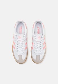 Bílé tenisky s růžovými akcenty, obsahující tři pruhy, šněrování a béžovou semišovou špičku. Gumová podrážka a polstrovaný límec.