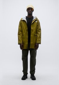 Αδιάβροχο μπουφάν ελιάς με γκρι επένδυση, μαύρο μπλουζάκι, παντελόνι ελιάς και μαύρα παπούτσια. Αξεσουάρ περιλαμβάνουν μπεζ σκουφάκι και γυαλιά ηλίου.