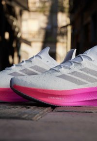 Scarpe da ginnastica bianche con accenti grigi, caratterizzate da una parte superiore testurizzata e una suola intermedia rosa brillante etichettata "LIGHT BOOST", posizionate su una superficie di legno.