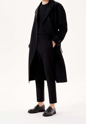 Homme portant un long trench-coat noir, un pantalon noir, une chemise noire et des mocassins en cuir noirs, debout avec la main dans la poche.