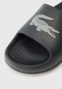 Svart halksandaler i gummi. Utrustad med öppen tå och strukturerad grepp-sula. Dekorerad med en grå krokodil-logotyp på ovansidan.