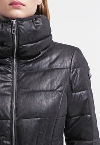 Schwarze Daunenjacke mit hohem Kragen, mit Reißverschluss, strukturiertem Stoff und horizontalen Nähten am Körper.