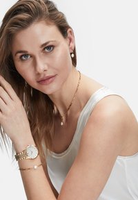 Guldtonet armbåndsur med metalrem, parret med en matchende guldlænke med en perleaccent, båret med en ærmeløs top.