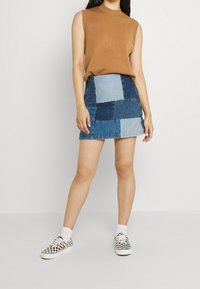 Spódnica z naszywkami z dżinsu w jasnych i ciemnych odcieniach niebieskiego, o dopasowanym kroju. W zestawie z beżowym, bezrękawnym topem i butami w kratkę.