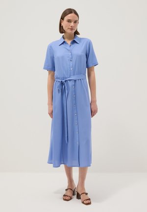 Vrouw draagt een lichtblauwe midi-jurk met korte mouwen en knopen, voorzien van een ceintuur in de taille en bruine sandalen met hak, staand op een witte vloer.