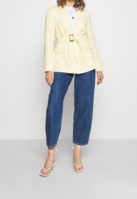 Light yellow blazer with a belted waist, two front pockets, and a notched collar, paired with loose blue jeans and strappy white heels.