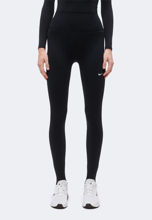 Person iført sorte høj-talje leggings med lille hvidt Nike-logo på låret og hvide Nike-løbesko.