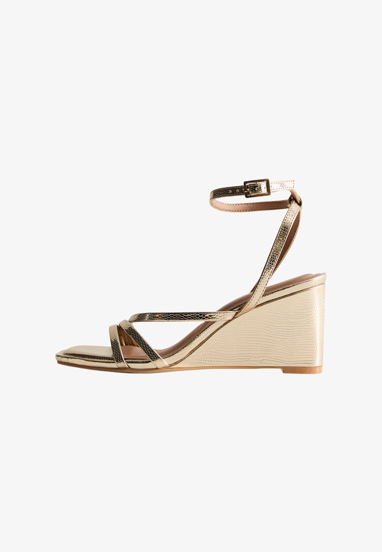 Gouden en beige sleehak sandalen met gekruiste banden, enkelbandje, gestructureerd faux leer en een ontwerp met sleehak. Gladde afwerking en lichte houten zool.