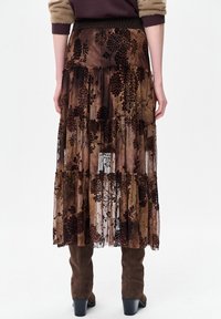 Lange, gelaagde rok van doorschijnende stof met bloemenpatronen in donkerbruin en beige tinten. Heeft een aangesloten tailleband en een semi-transparante afwerking.