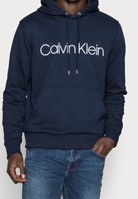 Man som bär en marinblå hoodie med vit "Calvin Klein"-text, kombinerad med blå jeans, den ena handen i fickan och den andra avslappnad.