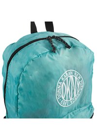 Mochila de nylon en color azul verdoso con cremalleras negras, que presenta un logo blanco circular y una superficie texturizada. Incluye dos correas de tela para llevar.