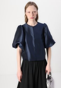 Marineblå satin topp med puffede ermer, rund halslinje og en detalj med frontseam. Kombinert med et flytende svart skjørt og holder en sølvveske.