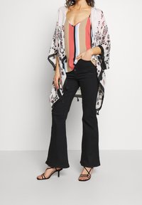 Kimono léger à motifs floraux blancs, bordé de noir avec des accents de pompons, porté sur un débardeur multicolore et un pantalon noir évasé.
