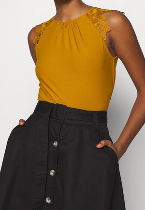 Mouwloze mosterdgele top met bloemrijke kantdetails op de schouders, ingestopt in een hoog getailleerde zwarte rok met knopen aan de voorkant en een riem.