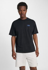 Zwarte katoenen t-shirt met korte mouwen, met een klein blauw logo op de borst. Gecombineerd met lichtgrijze shorts.