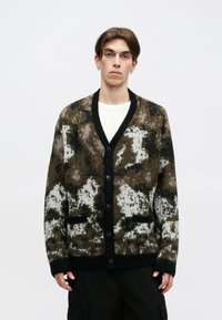 Pletena jakna s uzorkom u vojnom stilu u smešnim, crnim i belim tonovima. Ima V-izrez, dugmad za zatvaranje i dva prednja dzepa.